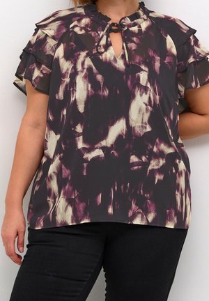 Woman wearing a short-sleeve, ruffled blouse with a black, cream, and purple abstract pattern and black pants against a white background.