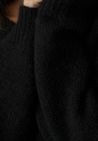 Pull en maille noire avec un tissu texturé et duveteux. Il dispose d'un col montant et de détails en côtes, offrant un aspect chaleureux et doux.