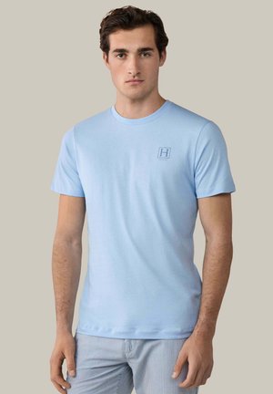 Mann trägt ein hellblaues kurzärmeliges T-Shirt mit kleinem "H"-Logo auf der Brust und helle gestreifte Hosen, steht vor einem einfarbigen Hintergrund.