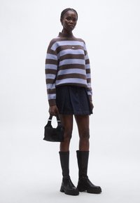 Lila és barna csíkos kötött pulóver magas nyakrésszel. Sötét farmer szoknyával, térdig érő fekete csizmával és kis fekete kézitáskával párosítva.