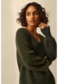 Donkergroene, gebreide oversized trui met een V-hals, ballonsleeves en een zachte textuur. Voorzien van geribbelde manchetten en een relaxte pasvorm.