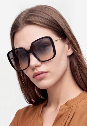 Žena s rovnými hnedými vlasmi, ktorá nosí veľké štvorcové čierne slnečné okuliare a hnedý top, pozerá mierne do strany.