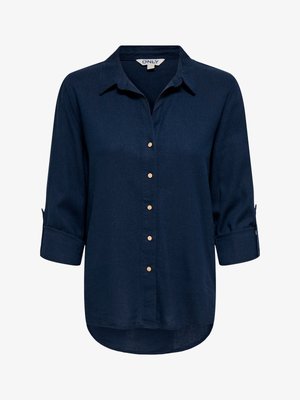 Blouse pour femme à manches longues bleu marine avec col, sept boutons beiges à l'avant, et manches retroussées maintenues par des pattes boutonnées.