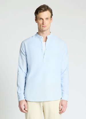 Homme aux cheveux courts et bruns portant une chemise bleu clair à manches longues avec un col mao et un pantalon beige sur un fond uni.