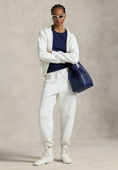 Γυναίκα που φοράει λευκό φούτερ με κουκούλα, λευκή φόρμα, λευκά αθλητικά παπούτσια και γυαλιά ηλίου σε σχήμα γάτας, κρατώντας μια μεγάλη τσάντα tote σε σκούρο μπλε χρώμα.