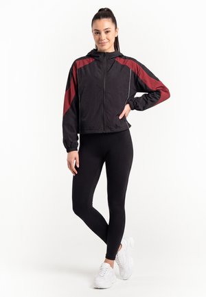 Giovane donna in piedi che indossa una giacca nera e rossa con zip, leggings neri e scarpe da ginnastica bianche su uno sfondo bianco semplice.