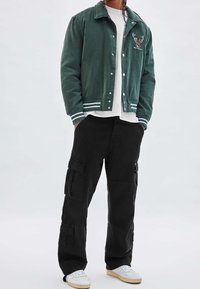 Grön kordfloret varsityjacka med broderad älgdesign, kombinerad med svarta cargobyxor med flera fickor och vita sneakers.