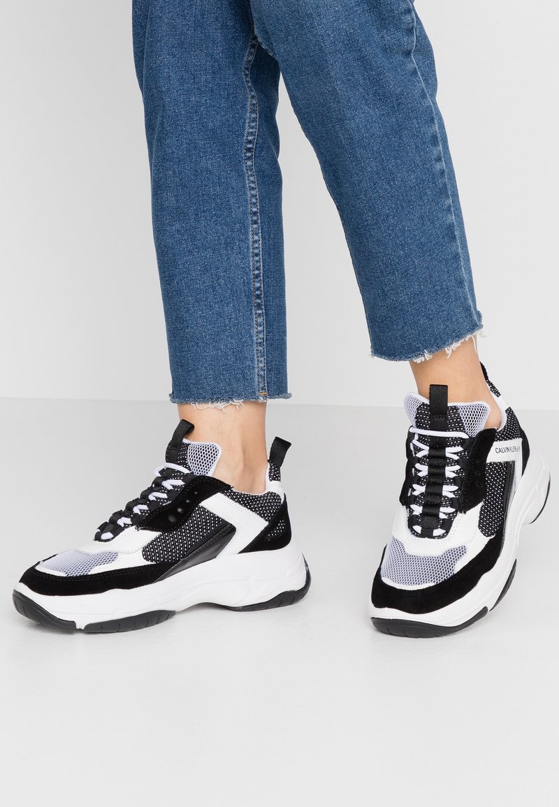 Baskets noires et blanches avec des textures en mesh et daim, une semelle épaisse, des lacets noirs et une languette de tirage à l'arrière. Portées avec un jean bleu raccourci.