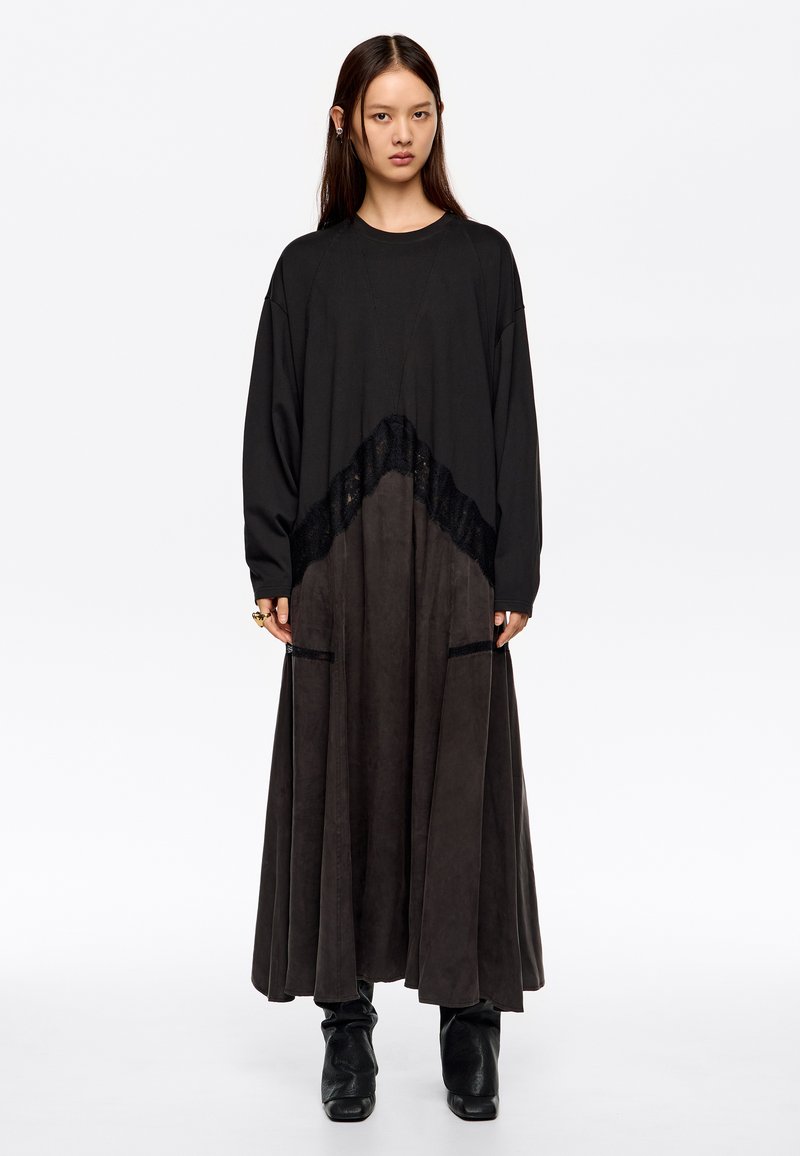 Długa czarna sukienka z fakturowanym detalem koronkowym w pasie. Spódnica ma luźną sylwetkę, wykonaną z miękkiego materiału o matowym wykończeniu.