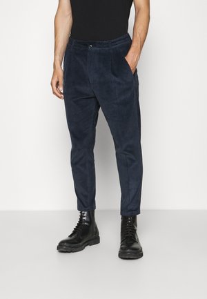Mann trägt dunkelblaue Cordhosen mit Seitentaschen und schwarze Schnürstiefel, steht vor einem einfarbigen hellen Hintergrund.