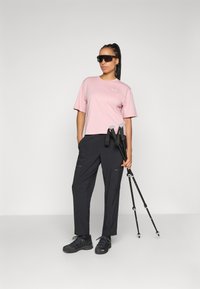 Helles rosa Kurzarmhemd mit kleinem Logo, kombiniert mit schwarzen Cargo-Hosen mit Reißverschlüssen und schwarzen Wanderschuhen, während er Trekkingstöcke hält.