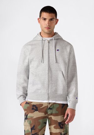 Junger Mann, der einen hellgrauen Kapuzenpullover mit Reißverschluss und Kordeln sowie Tarn-Cargohosen trägt und mit den Händen in den Taschen vor einem schlichten Hintergrund steht.