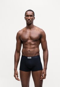 Boxer brev neri in cotone con banda logo bianca. Texture liscia, design aderente e gambe corte. Modello in piedi contro uno sfondo semplice.