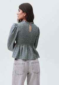 Blusa verde a fantasia con corpino arricciato, maniche a sbuffo e apertura a oblò sul retro. Abbinata a jeans in denim grigio chiaro.