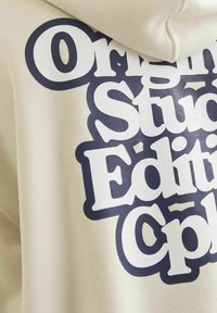 Felpa beige con grandi stampe di testo blu navy che dicono "Original Studio Edition Cph" sui lati, con una texture di tessuto morbido.