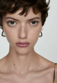 Close-up van een persoon met kort, donker haar, een gladde huid en minimale make-up. Ze dragen ronde, zilveren oorbellen tegen een neutrale achtergrond.
