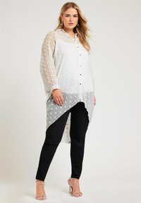 Túnica blanca transparente con lunares texturizados, dobladillo asimétrico, cierre de botones y mangas largas. Combinada con jeans skinny negros y tacones metálicos.