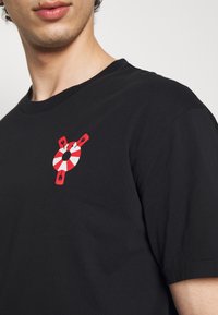 Černé bavlněné tričko s grafickým potiskem červeného a bílého záchranného kruhu na levé straně hrudi. Krátké rukávy a kulatý výstřih.