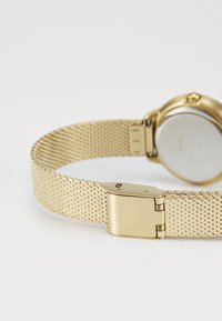 Bracelet de montre en maillage métallique doré avec un fermoir rectangulaire, un cadran de montre rond et un dos lisse argenté. Le design présente une esthétique minimaliste.