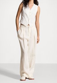 Žena v bezrukávové bílé košili na knoflíky, s vysokým pasem a širokými krémovými kalhotami a krémovými brogue botami, stojící s jednou rukou v kapse.