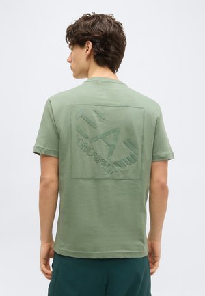 Νέος άνδρας με κοντά σγουρά μαλλιά που φοράει ανοιχτό πράσινο T-shirt με ανάγλυφο λογότυπο "EA7 Emporio Armani" στην πλάτη.