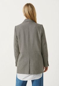 Blazer à motif pied-de-poule en noir et blanc, avec épaules structurées, deux fentes au dos, et une texture de tissu lisse.