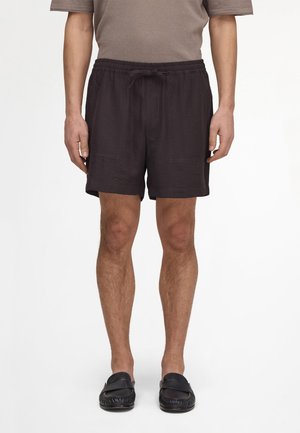Mand iført sorte shorts med snøre, brun kortærmet skjorte og sorte slip-on sko, stående mod en ensfarvet baggrund.