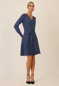 Abito in maglia blu con scollo a V e maniche lunghe, caratterizzato da un corpo aderente e una gonna svasata. Completo di cintura in vita annodata e texture liscia. Décolleté neri.
