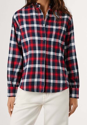 Kobieta ubrana w koszulę w czerwoną, granatową i białą kratę z długim rękawem, do tego jasnobeżowe spodnie, na jednolitym jasnym tle.