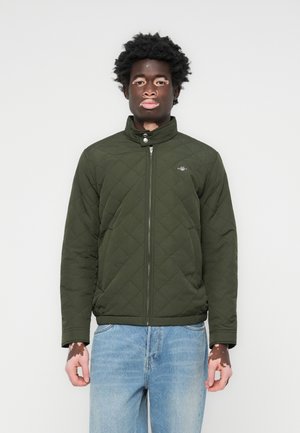 Quilted olive green jacket with a high collar, front zipper, and tonal logo. Paired with light blue jeans for a casual look.