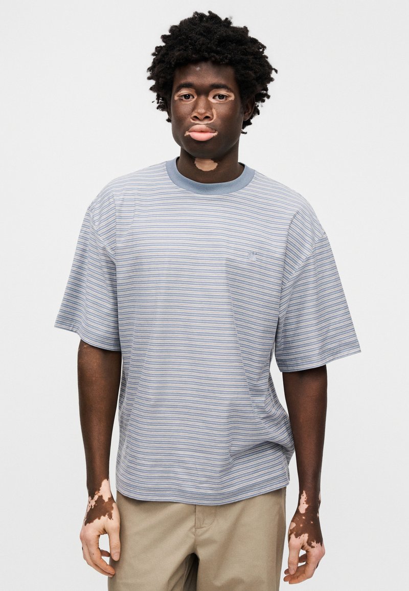 Striped t-shirt in light blue with gray, short sleeves, round neck, relaxed fit, subtle logo detail on chest, paired with beige pants.
