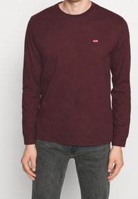 Burgundy långärmad skjorta i bomull med rund halsringning. Har en liten röd Levi's-logotyp på bröstet. Bärs med grå jeans.