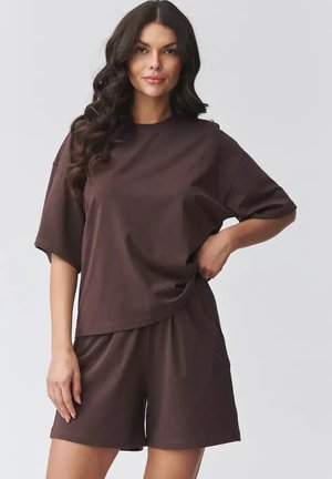 Kobieta z długimi falującymi włosami, ubrana w luźny ciemnobrązowy t-shirt i dopasowane szorty, stojąca z ręką na biodrze na jasnym tle.