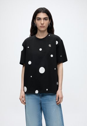 Frau mit langen dunklen Haaren, die ein schwarzes T-Shirt mit weißen Punkten und hellblaue Jeans trägt, steht vor einem schlichten Hintergrund.