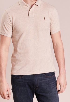 Mann trägt ein hellbeiges kurzärmeliges Poloshirt mit kleinem dunklem Logo auf der Brust und dunkelblaue Jeans, steht vor neutralem Hintergrund.