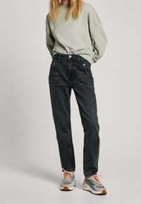 Jeans de mezclilla negra con un corte recto, bolsillos frontales y costuras sutiles, combinados con un sudadera verde claro y zapatillas multicolores.