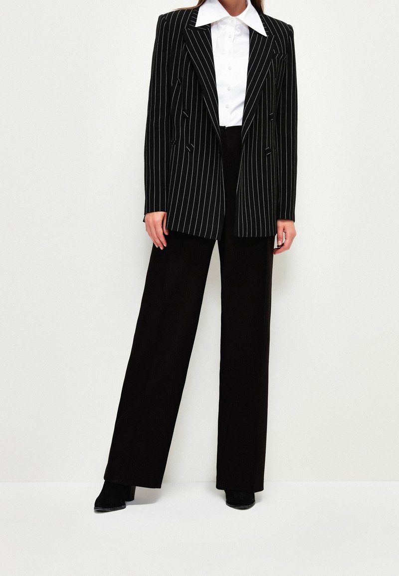 Nainen, joka on pukeutunut mustiin leveälahkeisiin housuihin, musta-valkoraidalliseen bleiseriin, valkoiseen napitettuun puseroon ja mustiin korkokenkiin.