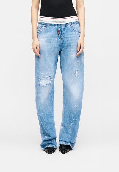 Άτομο που φοράει ανοιχτό μπλε φθαρμένα τζιν με φαρδιά μπατζάκια, μαύρα μυτερά παπούτσια και μαύρη μπλούζα, στέκεται μπροστά σε λευκό απλό φόντο.