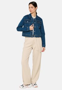 Vidutinio mėlynumo džinsinė striukė su trumpu dizainu, su dviem krūtinės kišenėmis, sidabriniais mygtukais ir kontrastine siuvimo linija. Suderinta su šviesiai kreminės spalvos kelnėmis.