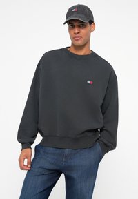 Felpa grigia scura con scollo a girocollo e polsini a coste, dotata di un piccolo patch con logo. Abbinata a jeans blu e a un cappellino coordinato.