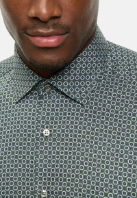 Hemden mit Knöpfen, aus grauem Stoff mit einem wiederholenden blauen und weißen Blumenmuster; verfügt über einen klassischen Kragen und zwei silberne Knöpfe.