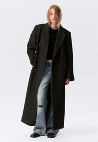Doppelreiher langer Mantel aus dunklem Fischgrätstoff, kombiniert mit einem schwarzen Croptop und ausgewaschenen hellblauen, weit geschnittenen Jeans. Schwarze Schuhe.