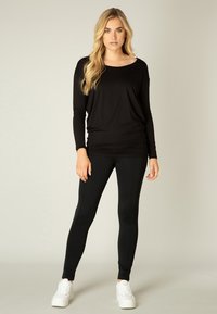 Zwarte lange mouw top met een ronde hals, gecombineerd met zwarte leggings. Het model draagt witte sneakers. Zacht materiaal met een ontspannen pasvorm.
