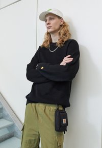 Schwarzer Strickpullover mit langen Ärmeln, grüne Cargo-Hosen und eine weiße Mütze. Verfügt über eine kleine schwarze Tasche, die an der Taille befestigt ist. Silberne Halskette.