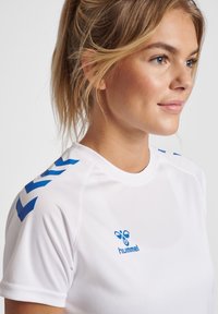 Hvid atletisk skjorte med blå detaljer og et logo. Skjorten har korte ærmer og en rund krave med chevronmønstre på skuldrene.