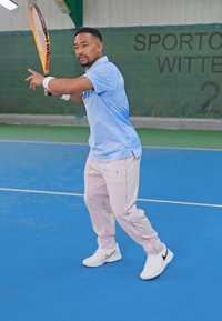 Tennisspiller i lyseblå skjorte og rosa bukser, som holder en oransje racket, på en blå innendørs bane med grønn bakgrunn.