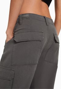 Primo piano di una persona che indossa pantaloni cargo grigi con tasche a toppa, una mano infilata nella tasca laterale e una vita visibile con un top nero.