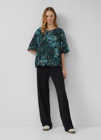 Blouse florale teal à manches courtes, avec un col rond et une coupe ample, associée à un pantalon noir à pattes larges et des chaussures de couleur claire.