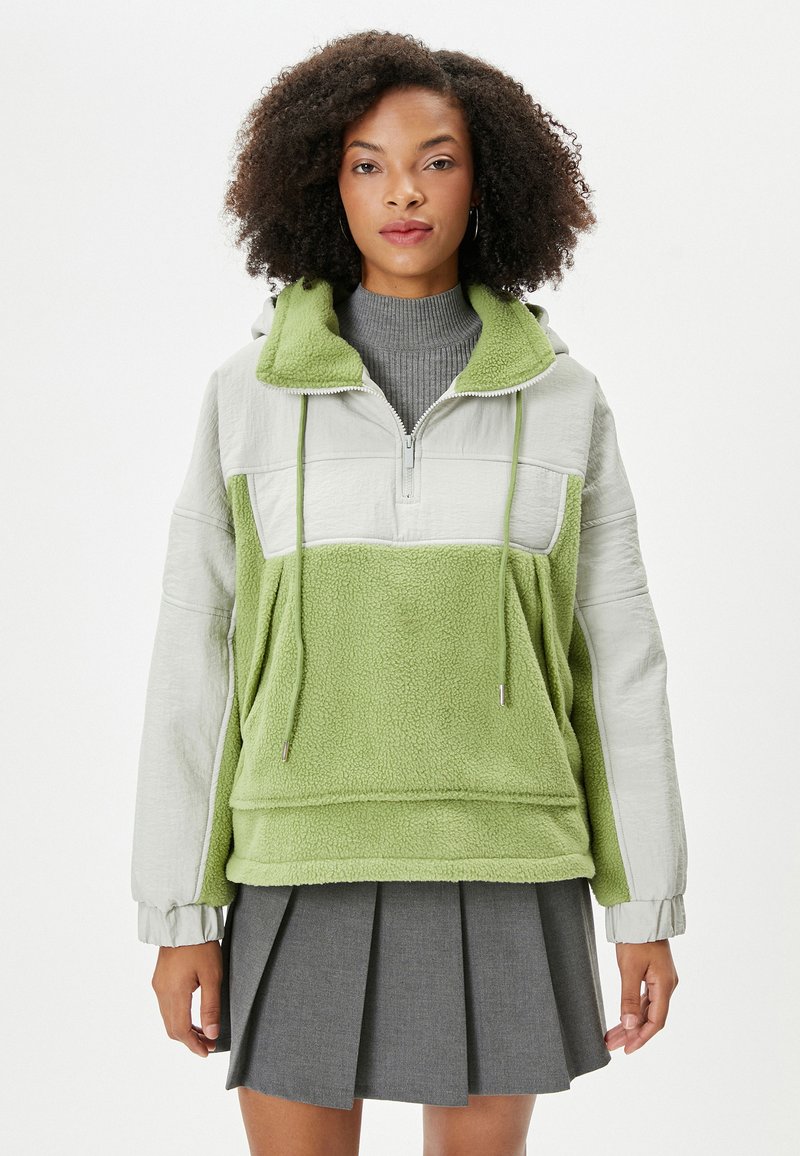 Chaqueta de forro polar verde con media cremallera, detalles en gris, cordones y un bolsillo frontal. Textura suave y corte relajado.