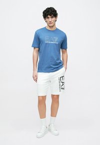 Μπλε μπλουζάκι με λευκό λογότυπο "EA7 Emporio Armani", συνδυασμένο με λευκά σορτς που έχουν μαύρο λογότυπο στο πλάι, φορεμένα με λευκά αθλητικά παπούτσια.